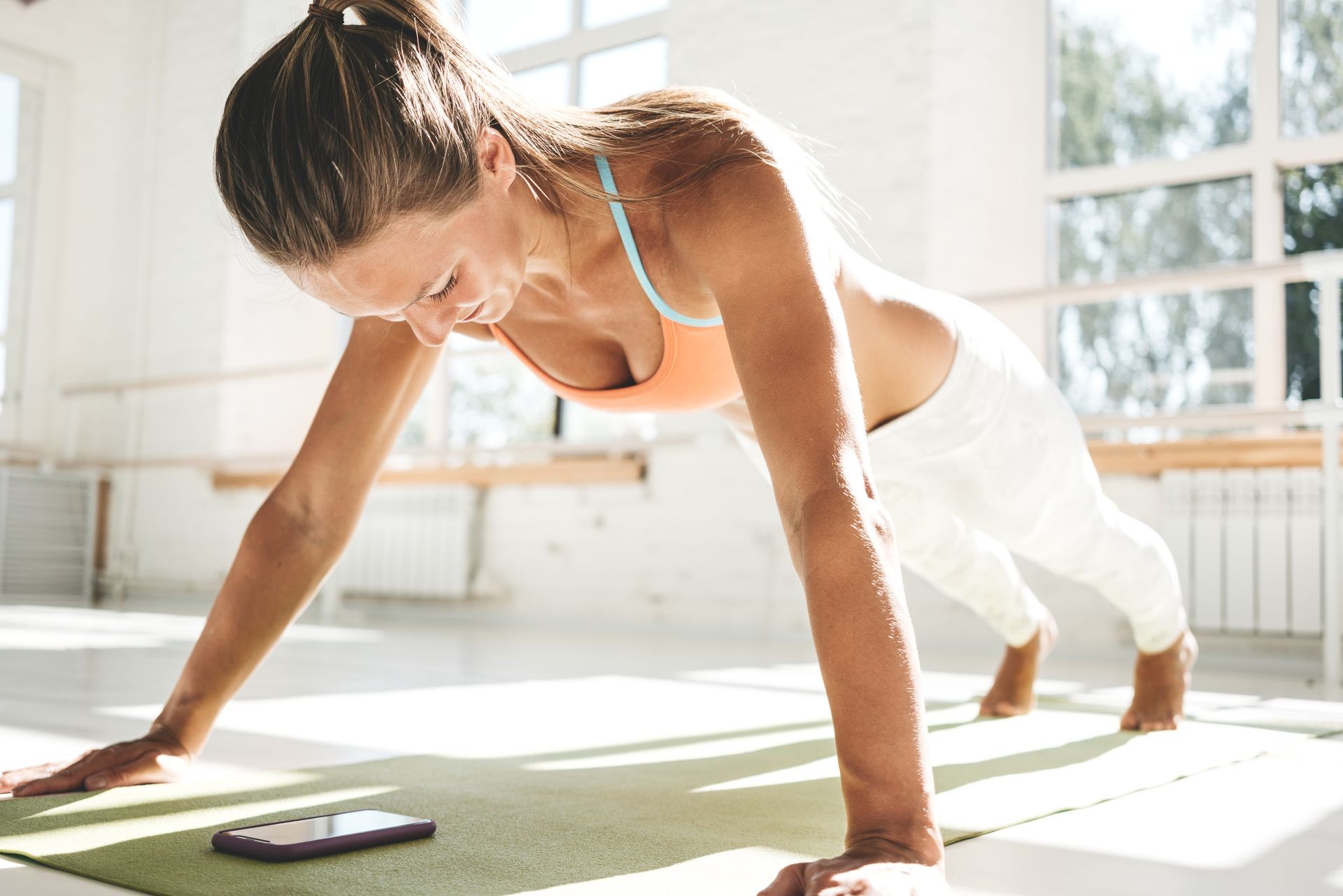 Sporty sunburn woman doing push-ups on fitness mat and using smartphone for counts results in fitness app. Female athlete exercising push up in sunny gym. Healthy lifestyle concept